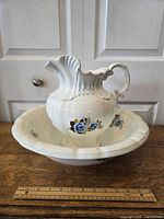 Side angle photo of white ceramic pitcher placed inside matching wash basin, both with blue floral decoration and scalloped edges. A ruler is placed in front showing approximate dimensions.