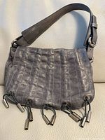 Front view of grey leather handbag showing ruched texture and silver tube hardware hanging from cords at bottom.