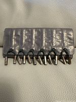Front view of the grey leather clutch purse showing vertical stitched lines and seven silver metal tube embellishments hanging from black cords.