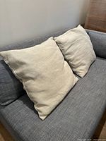 Pair of large off-white cotton and jute pillows resting on a grey couch, showing general size and appearance.