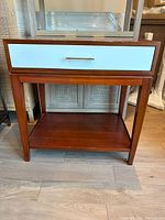 Front view of mid-century modern mahogany side table with white drawer and open shelf below, showing wood grain and metal drawer handle.