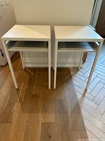 Front view of the pair of white metal end tables placed side by side on a wooden floor.