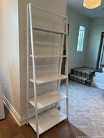 Full view of tall white metal folding bookcase against wall, showing five shelves and angled side supports.