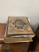Oblique angle view of thick antique Holy Bible showing leather cover with embossed cross and gold 'Holy Bible' lettering, visible wear and binding damage.
