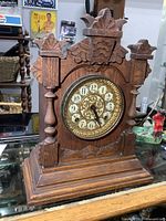 Front view showing ornate carved wooden mantle clock with decorative round dial and gold accents.