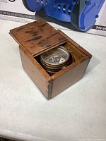 Antique brass compass in the wooden box with sliding lid partially open, showing the compass dial and gimbal mount inside the box.