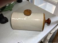 Photo showing the side view of the cylindrical stoneware foot warmer, cream-colored with brown glazed edges and wooden knob on top.