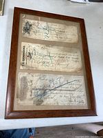 Three vertically mounted antique cheques with handwritten and printed details, cancellation and stamps, inside an oak frame.