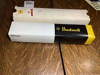 Original packaging of Bushnell riflescope showing white box with black and yellow Bushnell branding on wooden table.