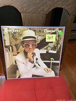 Front cover of the Elton John Greatest Hits LP showing Elton John in a white suit and hat with pink sunglasses, sitting at a piano.