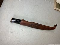 Vintage Normark fillet knife in brown leather sheath laying flat on table, showing exterior of sheath and partial handle.