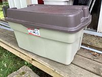 Side view of large Rubbermaid storage tote with brown hinged lid closed, placed on wooden surface outdoors.