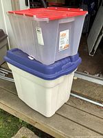 Two stacked plastic storage totes: top tote is HDX branded clear plastic with red lid, bottom tote is white Rubbermaid with blue lid.