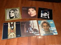 Photo of six vintage vinyl album covers on hardwood floor, including Olivia Newton John, Barbra Streisand, Irene Cara, Linda Ronstadt, Carole King.