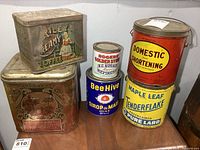 Front view of all six vintage kitchen tins arranged on wooden surface showing Riley's Creamy Toffee, Domestic Shortening, Maple Leaf Tenderflake and smaller blue, white, and red cans with aged surfaces