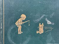 Close-up of front cover green cloth with gold embossed illustrations of Christopher Robin and Winnie the Pooh fishing.