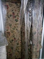 Photo showing the full couch wrapped in transparent plastic, floral upholstery visible with large flowers in cream, pink, red, and green, couch positioned vertically.