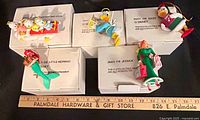 Five Disney ornaments displayed atop their labeled Grolier boxes with measuring ruler