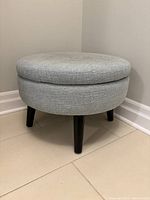 Front angle view of the light blue round fabric ottoman with three dark legs, showing the top and body upholstery clearly.