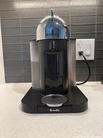 Front view of Breville Nespresso Vertuo machine showing black base, silver dome overhead, removable water tank on back left, and drip tray with metal grate.