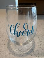 Close-up of a single clear stemless wine glass with blue "Cheers" print.
