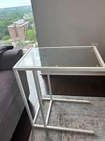 Side view of the white metal frame and clear glass top laptop stand positioned next to a grey sofa and a window.