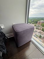 Side view of grey modern ottoman next to window, showing fabric texture and shape.