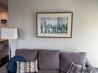 Wall-mounted framed abstract cityscape print above gray couch with pillows and nearby lamp.