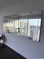 Full view of large rectangular mirror mounted on wall reflecting a cityscape and bedroom elements.