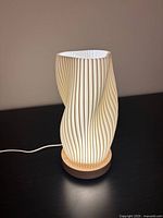 Front view of the LED lamp with sculptural twisted white shade on a wooden base, plugged in and lit