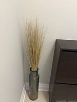 Full view of tall blue glass vase filled with natural-colored artificial reed grass, standing on floor against white wall.