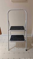 Full frontal view of white metal framed 2-step folding stool with black plastic/rubber steps on tile floor.