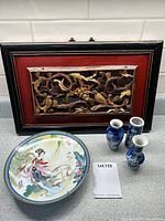 Framed carved wood panel with gilt floral carving details, red background and black frame; painted porcelain plate with colorful Asian figure; three blue and white floral miniature vases.