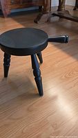 Black wooden milking stool with round seat and three turned legs, placed on a hardwood floor.