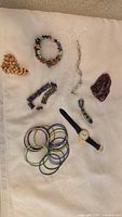 Lot items laid out on fabric including Bulova wristwatch, multicolor bangles, beaded bracelets, shell-like bracelet, and silver-tone chain and rhinestone bracelets.