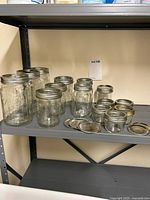 Shelf displaying 14 clear glass canning jars in mixed sizes with attached silver metal screw rings, alongside a group of loose silver and gold metal canning lids and rings.