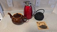 Brown ceramic teapot, red carafe, black cast iron teapot, and instruction card on countertop