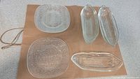 Set of six clear corn-shaped glass dishes and four textured square clear glass dessert plates arranged on brown paper surface.