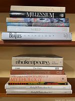 Photo showing the titles and spines of 13 hardcover books arranged in two stacks on wooden shelves, including Alistair Cooke's America, Millennium, The Beatles Anthology, Shakespeare works, and mythology titles.
