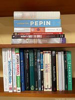 Photo showing stacked cookbooks on a shelf with titles visible including 'Essential Pepin', 'Cooking Italian', 'Weber's Way to Grill', 'The Food of China', and others.