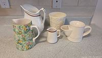 Five ceramic pitchers and jugs on a kitchen counter. Various sizes and designs. One floral multicolor green base pitcher, one large white pitcher with brown rim, one white pitcher with floral embossing, one white cup with circular pattern, one small creamer jug.