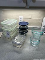 View of 11 assorted glass food storage containers displayed on counter. Includes containers with plastic lids in green trim and blue, and containers without lids.