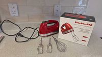 KitchenAid empire red hand mixer with power cord plugged out, two turbo beaters and one whisk displayed alongside the original box showing product details.