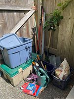 Photo showing assortment of gardening tools including shovels, rakes, hand tools, watering can, garden gloves, plastic storage bins, wire mesh pieces, and bags of supplies.