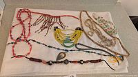 Beaded necklaces of various colors and materials laid flat on white cloth, showing 8-10 necklaces with beads of wood, glass, plastic, and shell in multiple colors and styles.