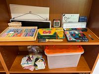 Shelf with art supplies including pastels, crayons, watercolors, markers, art boards, paint brush, fabric pieces, and storage tote.