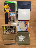 Top view of all items neatly arranged on the floor including file folders, staplers, hole punch, binder, clips, Kodak photo paper, poly folders, and labels.