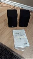Pair of black PSB Alpha LR1 bookshelf speakers with front view visible and owner's manual placed in front.