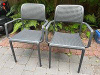 Front view of two dark gray plastic stackable outdoor patio chairs with perforated seats and backrests, armrests, placed on stone patio with fern plants in background.