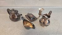 Front view of all five silver plated items displayed on a countertop including teapot, pitcher, chalice, gravy boat, and butter dish with lid.